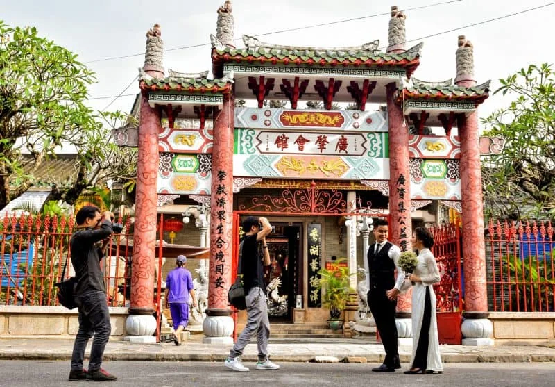 A couple poses for a wedding photoshoot in front of the ornate entrance gate of the Cantonese Assembly Hall Hoi An.
