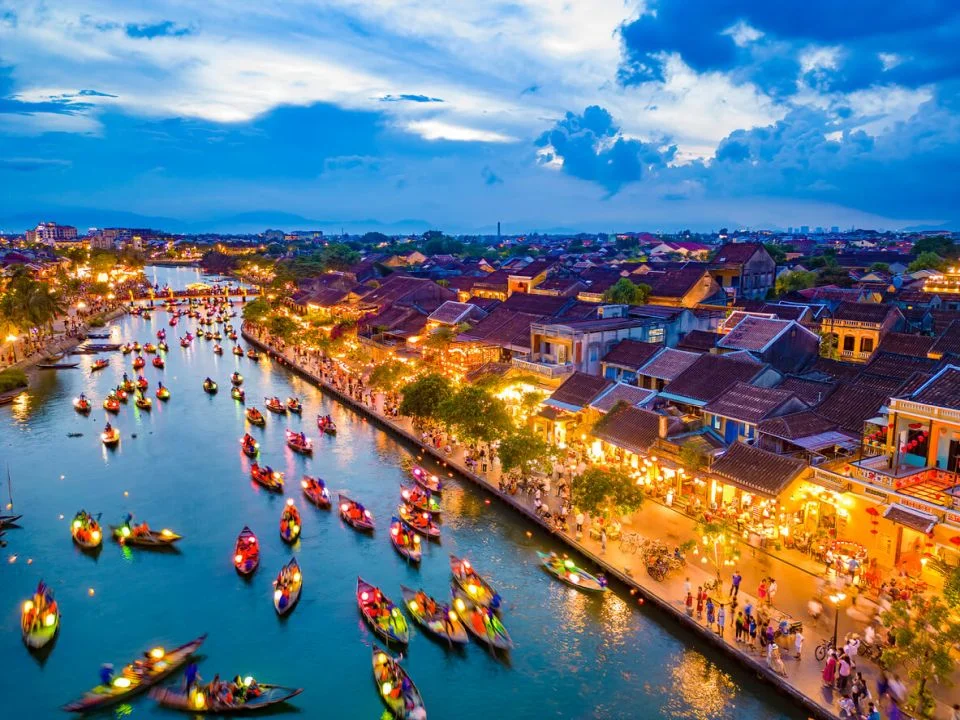 an aerial view of many colorful boats on the Hoai River during Hoi An Lantern Festival boat rides, with the illuminated ancient town on the banks at dusk.