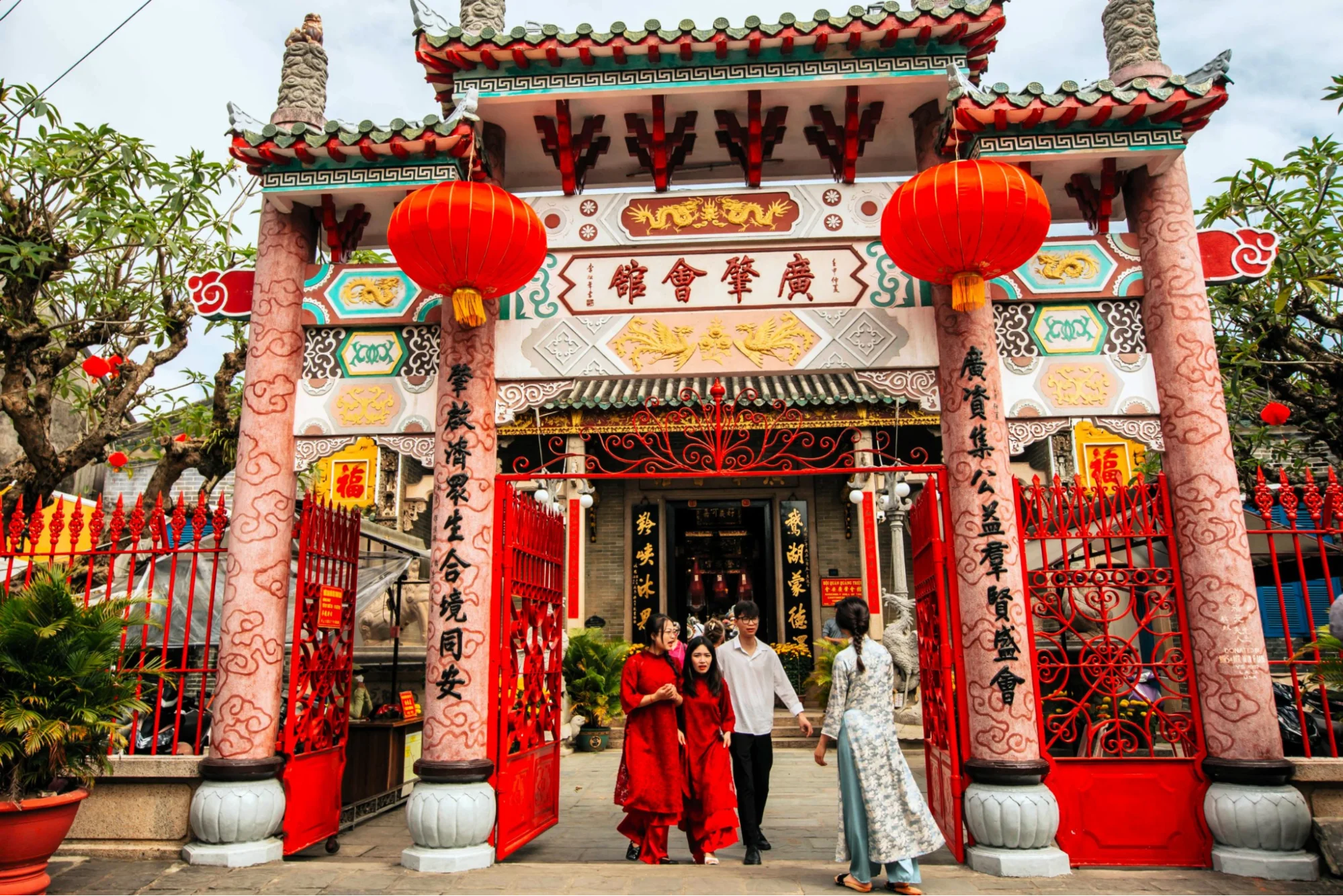 visitors walk through the vibrant, ornate entrance gate of the Cantonese Assembly Hall Hoi An, adorned with red lanterns.