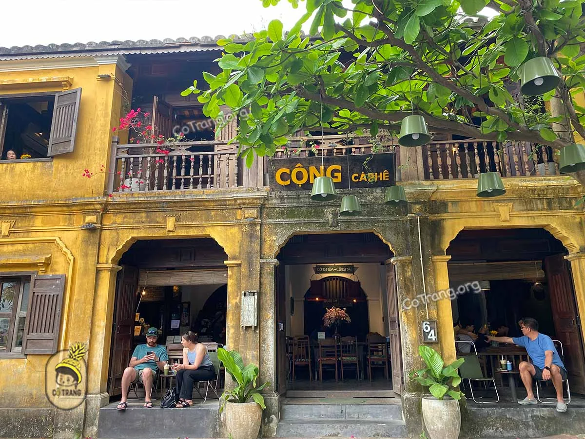 A sunny view of a traditional yellow French colonial coffee shop in Hoi An, Vietnam, decorated with colorful silk lanterns and lush bougainvillea flowers.