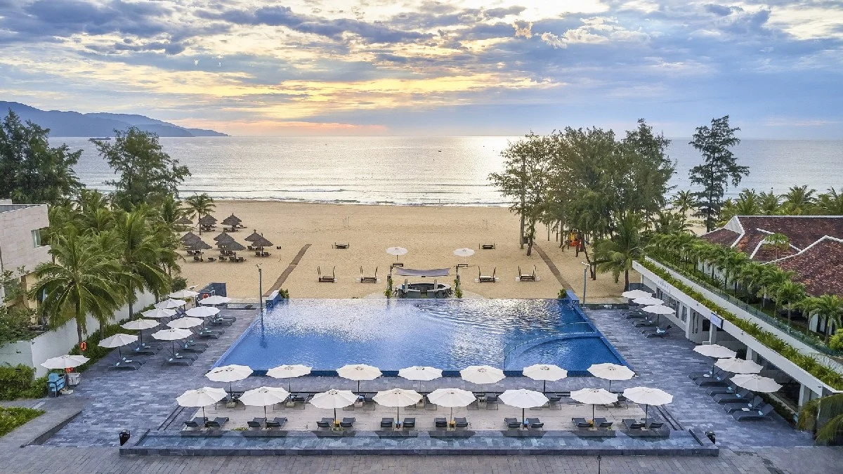 An aerial view of a luxurious beachfront resort pool and sandy beach at sunset, highlighting the modern amenities discussed in the Da Nang vs Hoi An debate.
