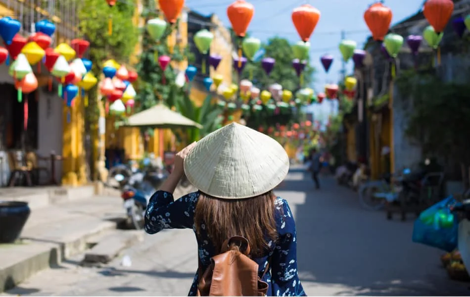 A person wearing a conical hat walks through a charming street in Hoi An, decorated with colorful lanterns, capturing the essence of Hoi An vs Hue.