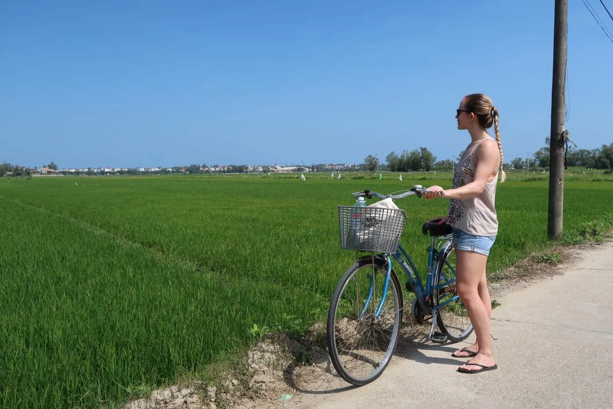 Woman pausing with a bicycle beside green rice paddies on a Hoi An cycling route.