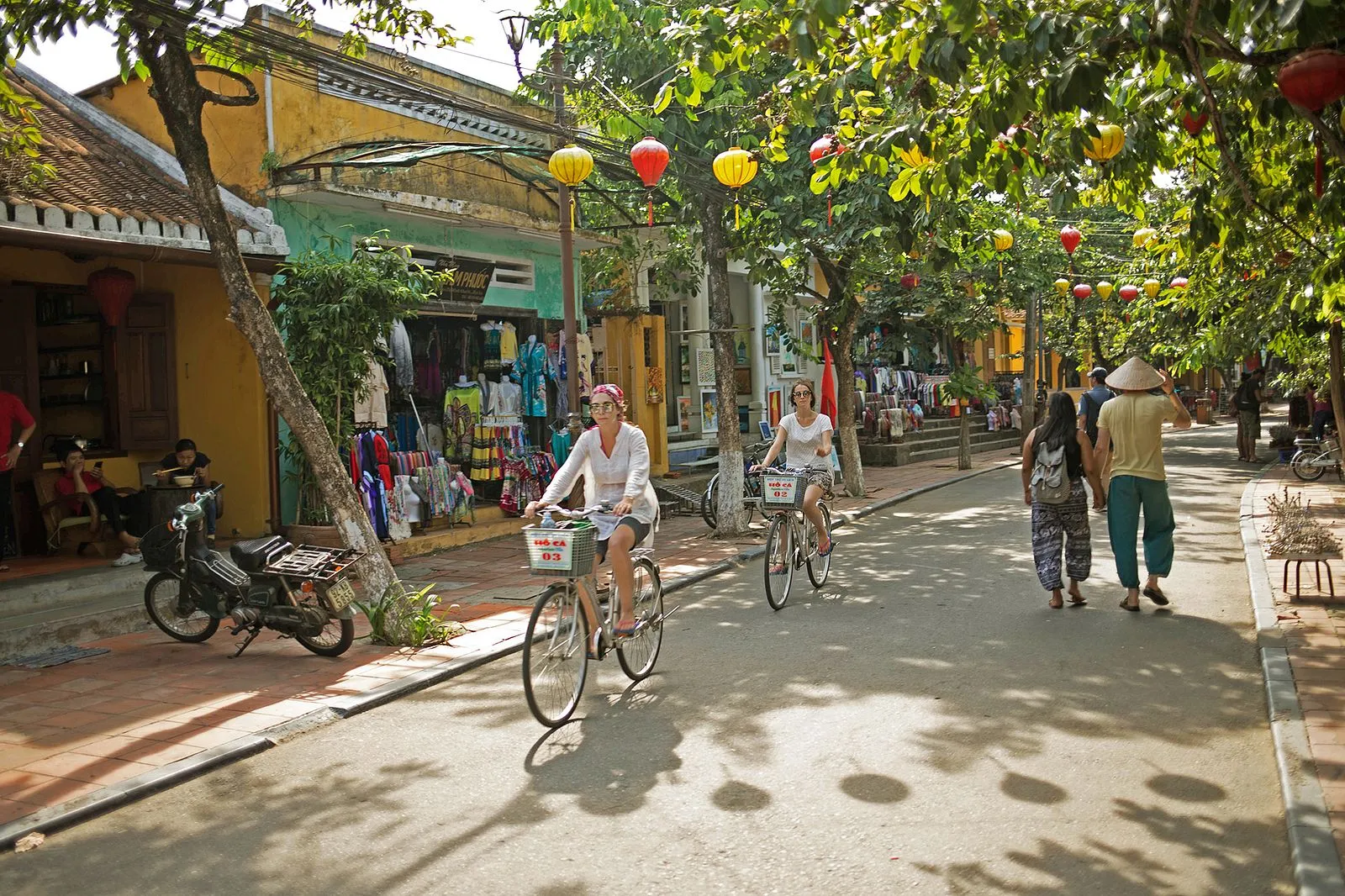 Two women riding bicycles under colorful lanterns on a paved Hoi An cycling route.