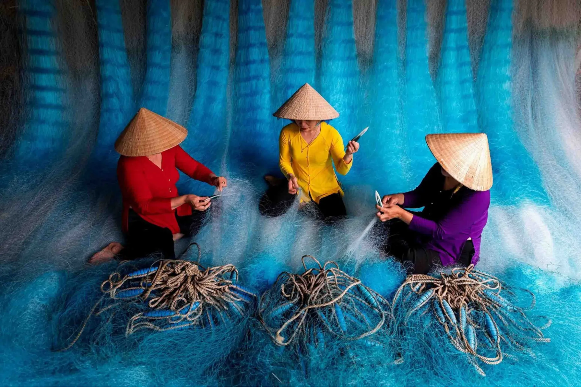 Women in conical hats mending large blue nets in a Hoi An fishing village.