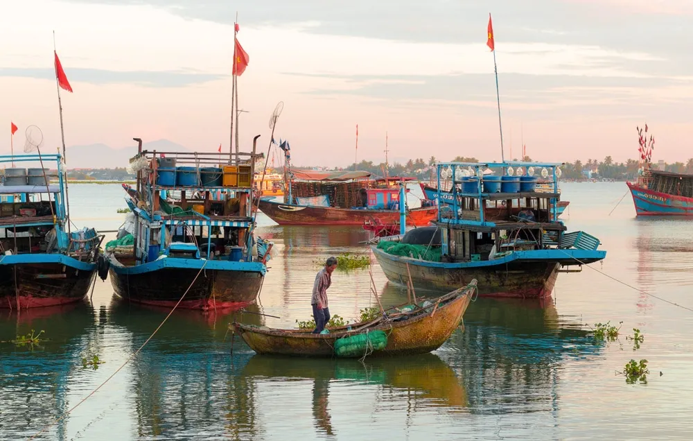 Fisherman on a small boat near colorful trawlers at a Hoi An fishing village.
