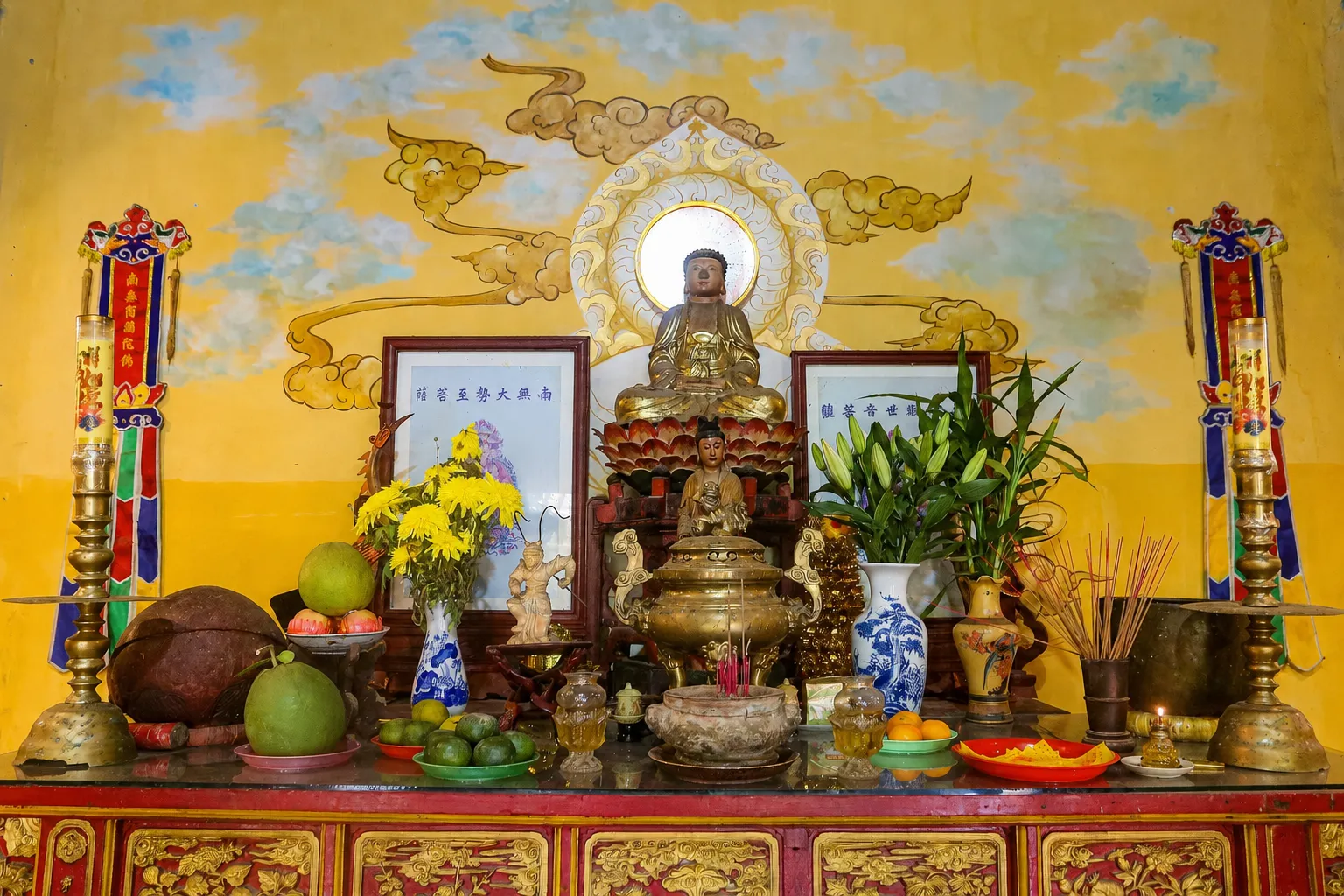 Buddhist altar with seated statues, incense burner, fresh flowers, and fruit offerings at quan am pagoda hoi an