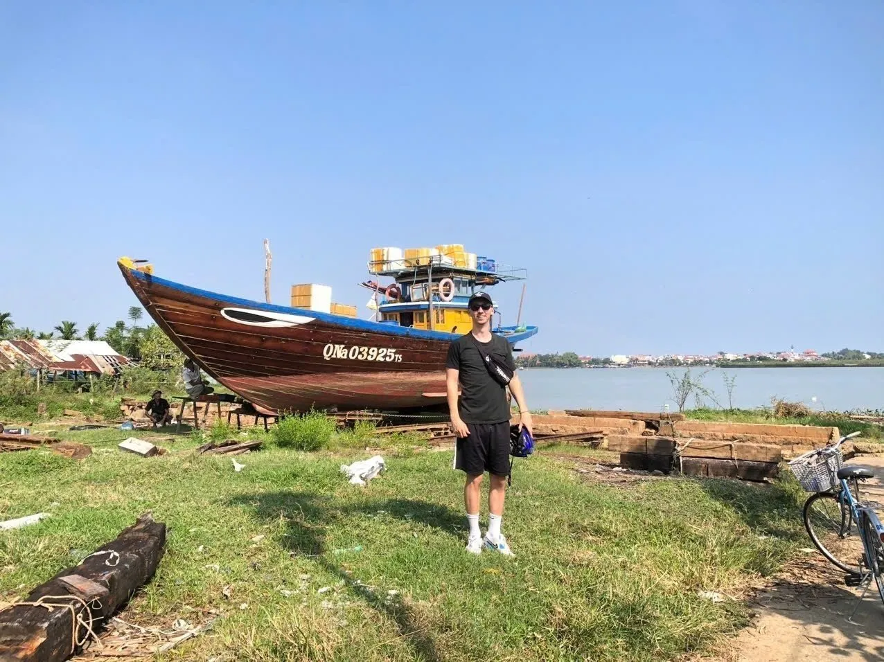 A visitor stands near a shipyard on Cam Kim Island, with a boat and the Thu Bon River Hoi An.